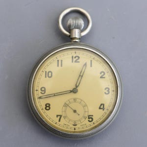 Può includere: Un orologio da tasca argentato con quadrante color crema e numeri neri. L'orologio ha un piccolo quadrante dei secondi e una corona strutturata. Il quadrante recita "SWISS MADE".