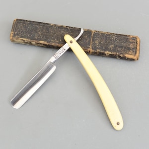 May include: A straight razor with a cream-coloured handle and a silver blade. The blade is marked with the text "GARANTIE SOLINGEN". The razor is next to a dark brown leather case.