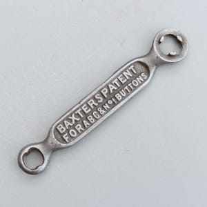 Puede incluir: Una herramienta metálica plateada con el texto "BAXTER'S PATENT FOR ABC & N°1 BUTTONS" en relieve. Tiene un lazo en cada extremo y una abertura circular dentada. La herramienta es probablemente vintage.