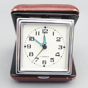 Peut inclure: Un réveil argenté avec un cadran blanc et des chiffres noirs. L'horloge est dans un étui en cuir marron et porte l'inscription "11 jewels".