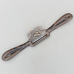 May include: A vintage drawknife with a metal blade and dark, worn wooden handles. The tool has a central blade housing with a screw. The handles have holes for hanging. This woodworking tool is designed for shaping wood.