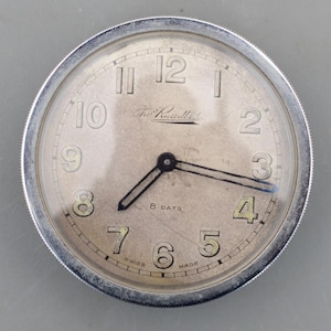 Può includere: Un orologio rotondo argentato con un quadrante chiaro. Presenta lancette nere, grandi numeri neri e le parole "8 DAYS" e "SWISS MADE". Il quadrante dell'orologio mostra anche il nome "Tho. Russell & Sons".