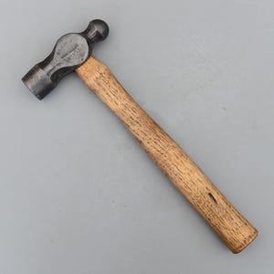 以下が含まれることがあります： 暗い金属製のヘッドと木製のハンドルが付いたボールピンハンマー。ハンマーヘッドは、片側に丸みを帯びた打撃面があり、もう片側にボール型のピンがあります。ハンドルは明るい茶色の木材で作られています。