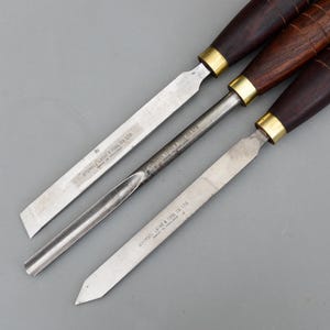 May include: Three wood turning chisels with dark brown handles, brass ferrules, and silver blades. The blades have the text "ARUNDEL LATHE & TOOL CO LTD. MADE IN ENGLAND". The chisels are arranged diagonally.