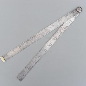 Può includere: Un antico righello pieghevole in metallo, con segni incisi per i pollici. Il righello è aperto, mostrando misure fino a 58.4 cm. Il metallo ha un aspetto usurato e invecchiato, con un terminale color ottone e un rivetto sulla cerniera.