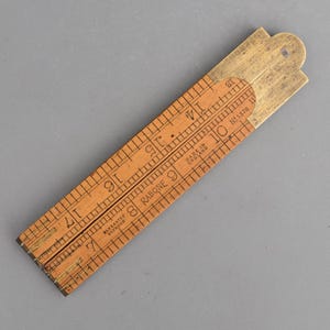 May include: A folding boxwood ruler with brass hinges and fittings. The light brown wooden segments feature black printed imperial measurement markings and numbers. Text on the surface reads: Warranted Boxwood, Rabone, No 1378, Made in England.