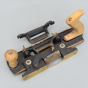 May include: A vintage woodworking plane with a wooden handle and knob. The plane is made of black metal with brass accents. The detached part has the number "30" on it.