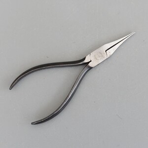 May include: A pair of needle-nose pliers with a black handle and silver-colored head. The pliers have a long, tapered nose and curved handles for gripping. The tool is designed for precision work and is likely used in jewelry making or electronics.