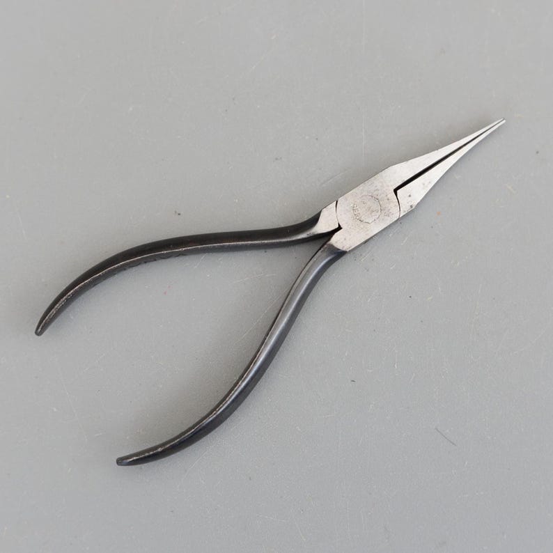 May include: A pair of needle-nose pliers with a black handle and silver-colored head. The pliers have a long, tapered nose and curved handles for gripping. The tool is designed for precision work and is likely used in jewelry making or electronics.