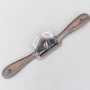 May include: A vintage drawknife with a dark, weathered appearance. The tool features a curved blade and two wooden handles, showing signs of age and use. The metal blade is a dark gray color, and the handles are a dark brown.