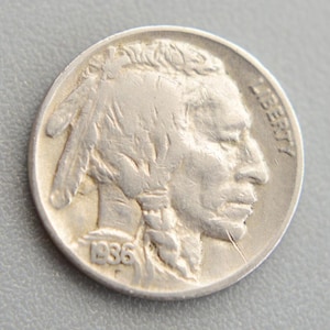 Puede incluir: Una moneda de níquel Buffalo de plata de 1936. El anverso presenta un perfil nativo americano con la palabra "LIBERTY" y la fecha. La moneda es redonda y tiene un aspecto desgastado.