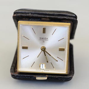 Puede incluir: Un reloj calendario Swiza vintage con una esfera cuadrada dorada y una esfera plateada. El reloj presenta agujas e indicadores dorados, y una pantalla de fecha en la parte inferior. El reloj está encerrado en un estuche de viaje de cuero negro.