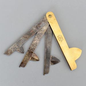 Może przedstawiać: Antyczna składana linijka z mosiądzu i stali, częściowo otwarta. Linijka ma mosiężny uchwyt i kilka stalowych ostrzy. Ostrza są oznaczone tekstem. Linijka leży na szarej powierzchni.