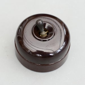 May include: A vintage-style, round light switch in a rich, dark brown color. The switch features a toggle lever and a circular base. The design is reminiscent of classic electrical fixtures, offering a blend of functionality and retro aesthetics.