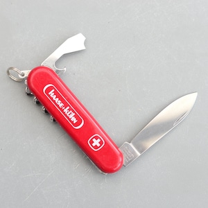 May include: A red pocket knife with a white Swiss cross and the text "Hase + Kuhn" on the handle. The knife has a blade, a bottle opener, a corkscrew, and a small screwdriver.