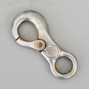 May include: A silver-coloured metal snap hook with a spring-loaded gate and two circular openings. The hook has a patent marking and the text "KUNGSÖRS YACHTYARD" stamped on its surface. Designed for securing ropes or other items.