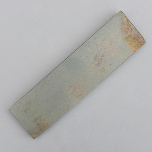 May include: A rectangular sharpening stone, approximately 15 cm long, with a grey-green colour. The stone has a slightly rough texture and a worn appearance, with a lighter, tan-coloured area at one end. Used for sharpening blades.