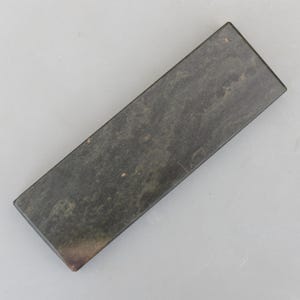 May include: A dark grey rectangular sharpening stone. The stone has a textured surface and is likely made from a dense material. It is used for sharpening tools, with dimensions of approximately 15 cm in length.