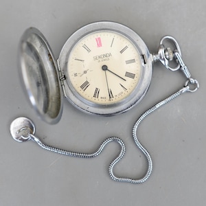 May include: A vintage Sekonda pocket watch with a silver case and chain. The watch face is cream-coloured with black Roman numerals and hands. The watch is open, revealing the time. The watch is marked "Sekonda 18 Jewels Made in USSR".