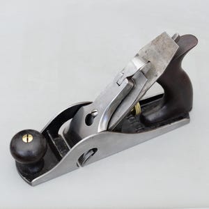 May include: A metal hand plane for woodworking featuring a silver steel body, a dark brown wooden handle and front knob, and a polished steel blade assembly. The tool includes brass adjustment screws and a lever cap mechanism for securing the blade.