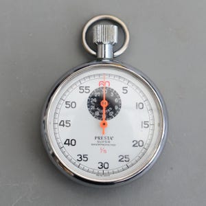 Könnte beinhalten: Eine silberfarbene Stoppuhr mit weißem Zifferblatt, schwarzen Zahlen und einem orangefarbenen Sekundenzeiger. Die Uhr trägt den Text "PRESTA SUPER SHOCKPROTECTED" und ist mit "SWISS MADE" gekennzeichnet. Sie hat einen Ring oben.