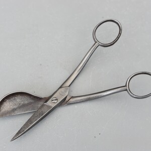 Può includere: Un paio di forbici vintage in metallo con una lama curva e un piccolo manico arrotondato. Le forbici sono in metallo scuro invecchiato e hanno un aspetto leggermente arrugginito.