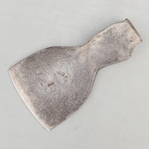 May include: A vintage, weathered metal axe head with a curved blade and a slightly curved handle end. The axe head has a worn, textured surface and a small, stamped marking near the center. The overall shape is asymmetrical.