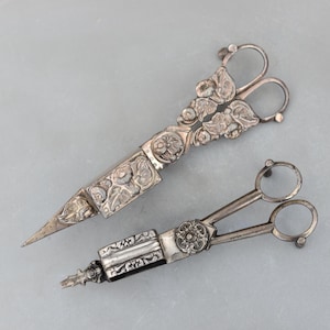 Puede incluir: Dos herramientas de costura antiguas de plata. Una es un par de tijeras con diseños florales y de hojas ornamentados en las asas y la carcasa de la hoja. La otra es una pequeña herramienta plateada con un asa circular y un centro decorativo.