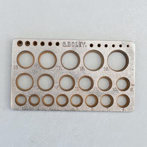 May include: A silver metal ring sizer with numbered holes for sizing rings from 5 to 19. The sizer is labelled "G.BOLEY."