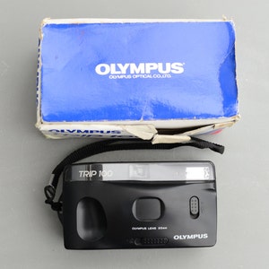 Könnte beinhalten: Eine schwarze Olympus Trip 100 35mm Filmkamera mit einem schwarzen Gurt. Die Kamera hat ein silbernes Objektiv und einen silbernen Blitz. Die Kamera befindet sich in einer Schachtel mit dem Olympus-Logo.