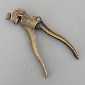 May include: A vintage brass saw set tool with a curved handle and adjustable head. The tool has a textured finish and a screw for adjustments. The overall design is compact and appears to be well-worn.
