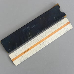 May include: A vintage slide rule with a wooden and paper construction, presented with a dark blue case. The slide rule displays measurements in inches. The text "ARMSTRONG" is visible. The case shows signs of wear.