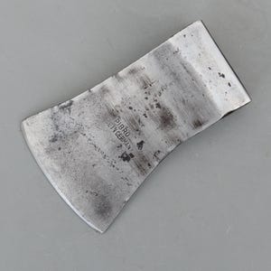 May include: A vintage axe head with a curved blade and a worn, metallic finish. The axe head has the text "PIBRO" and "MADE IN GERMANY" stamped on its surface. The axe head is a tool for chopping wood.