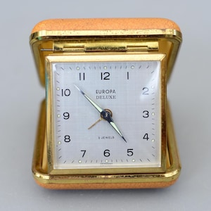 以下が含まれることがあります： 金色のケースと明るい文字盤を備えた、ヴィンテージのEuropa Deluxeトラベルクロック。文字盤には黒い数字と針があり、「EUROPA DELUXE」と「2 JEWELS」の文字が印刷されています。時計はタン色の合皮ケースに入っています。
