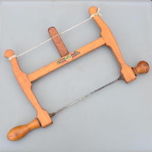 May include: A vintage wooden coping saw with a light brown finish. The saw features a thin, metal blade and a wooden handle. The frame is made of wood and has a string for tension. The saw has a label that reads "MARPLES & SONS".