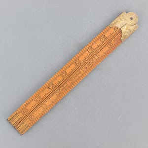 Può includere: Un righello pieghevole in legno vintage, con misure in centimetri da 1 a 66. Il righello è di colore arancione chiaro con segni neri e una cerniera in ottone. I testi "RABONE" e "BOYWOOD" sono visibili.