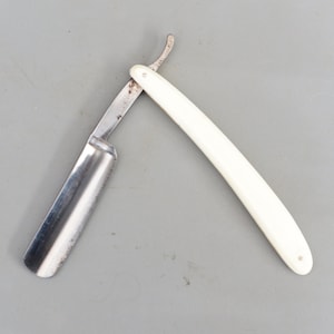 Peut inclure: Un rasoir droit vintage avec un manche en plastique blanc. Le rasoir a une lame en métal et une pointe arrondie.