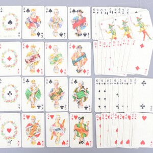 Może przedstawiać: Talia kart do gry z ozdobnymi ilustracjami postaci historycznych i motywów kwiatowych. Karty przedstawiają piki, karo, trefl i kier, z figurami i kartami numerowanymi. Karty są ułożone na szarej powierzchni.