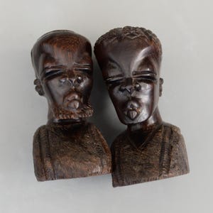 May include: Two dark brown wooden sculptures of human busts. The sculptures have detailed facial features, including closed eyes and open mouths. The busts are carved with textured clothing and are set against a neutral background.