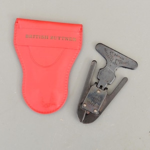 Peut inclure: Un outil métallique vintage avec une poignée en forme de T et des lames coupantes, à côté d'une pochette en cuir rouge. L'outil porte l'inscription "ST. BRITAIN HARDENED STEEL EDGES". La pochette est estampée "BRITISH BUTTNER".