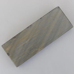 Puede incluir: Una piedra de afilar rectangular con una superficie gris y marrón moteada. La piedra está diseñada para afilar cuchillos y otras herramientas con filo. La piedra tiene una forma rectangular sólida.