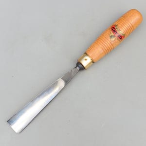 Può includere: Uno scalpello per intaglio del legno con una lama in acciaio lucido e un manico in legno. L'impugnatura ha una ghiera in ottone e un'etichetta rossa e oro con la scritta "TIRANTI". Lo scalpello è progettato per la lavorazione e l'intaglio del legno.