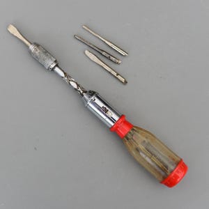 May include: A vintage multi-bit screwdriver with a clear plastic handle and a red base. The metal shaft has a twisted design. Three additional screwdriver bits are included.