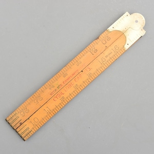 Peut inclure: Une règle pliante en bois vintage, avec des mesures en centimètres. La règle est marron clair avec des marquages et du texte noirs, dont "CYRIL LORD CAPETS." La règle est partiellement ouverte, montrant le mécanisme de pliage.