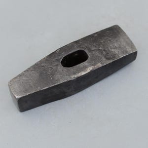May include: A dark gray, rectangular metal hammer head with a rectangular hole in the center. The hammer head has a slightly angled striking face and is designed for use with a handle. The tool is likely used for construction or metalworking.