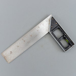 Puede incluir: Una escuadra de metal plateado con un nivel negro y una regla marcada en pulgadas. La regla está marcada con el texto "Stanley 42" y "Made in England".