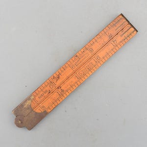 Könnte beinhalten: Ein orangefarbener, zusammenklappbarer Holzlineal mit schwarzen Markierungen und Text. Das Lineal ist mit Zahlen von 13 bis 18 Zoll (ca. 33 bis 46 cm) markiert. Der Text auf dem Lineal lautet "J. Rabone & Sons, Birmingham" und "Warrent Boxwood".