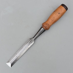 May include: A wood chisel featuring a long, rectangular steel blade with a beveled cutting edge and a cylindrical wooden handle. The metal shank connects the blade to the handle, which has a dark, reinforced cap at the end of the grip.