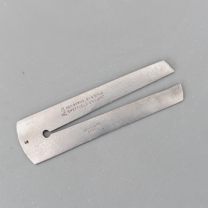 Puede incluir: Una herramienta de acero inoxidable con un diseño dividido, con el texto "GEO BARNS, EY & SONS SHEFFIELD ENGLAND" y "STAINLESS STEEL". La herramienta tiene un pequeño agujero y un extremo curvado, sobre una superficie gris.