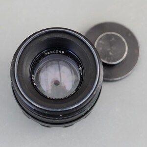 Op de afbeelding: Een zwarte cameralens met het nummer 7690046 aan de zijkant. De lens heeft een glazen element en een zwarte dop ernaast. De lens is ontworpen voor fotografie.